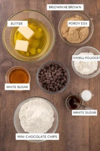 An overhead flat lay of baking ingredients for chocolate chip cookies, including bowls of flour, sugar, melted butter, eggs, and a pile of mini chocolate chips, arranged neatly on a light wooden table.